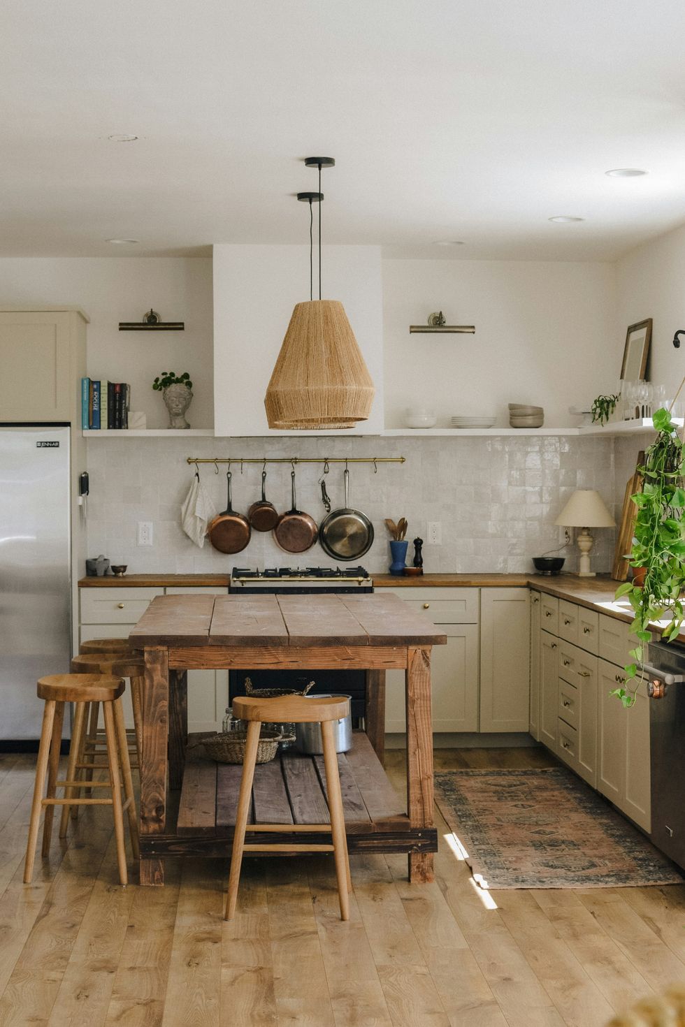 Rustic kitchen with wooden island, stools, hanging pans, and a woven pendant light.