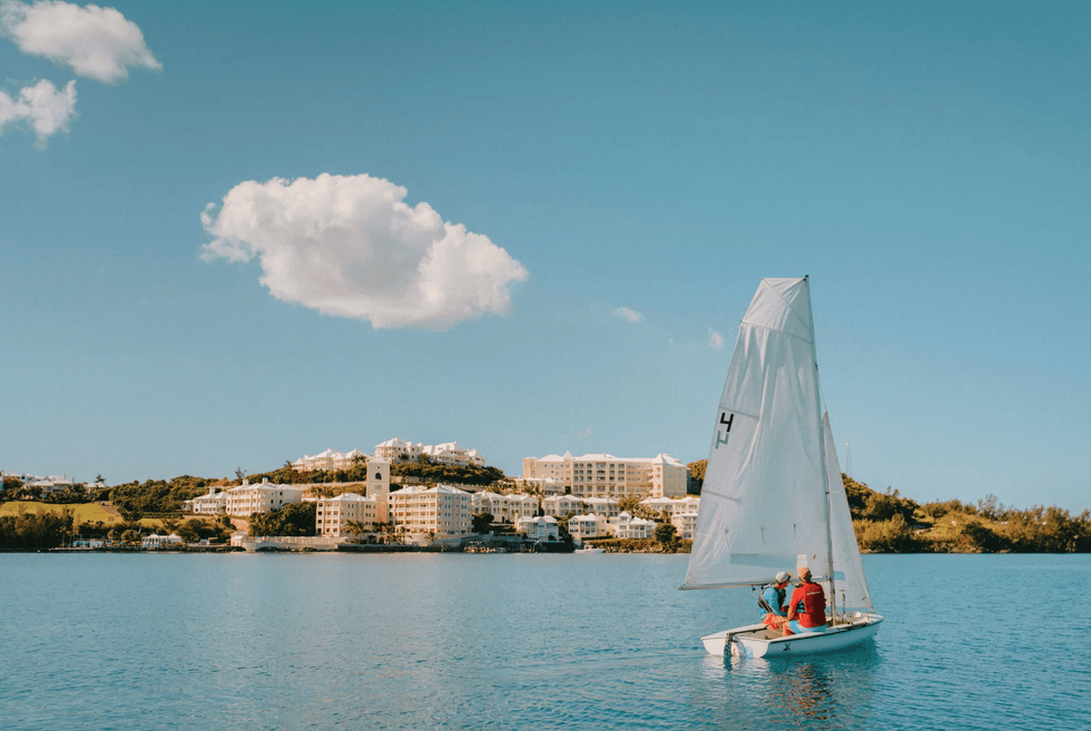 Sailboat on a blue sea with hilltop buildings and a fluffy cloud in the sky.