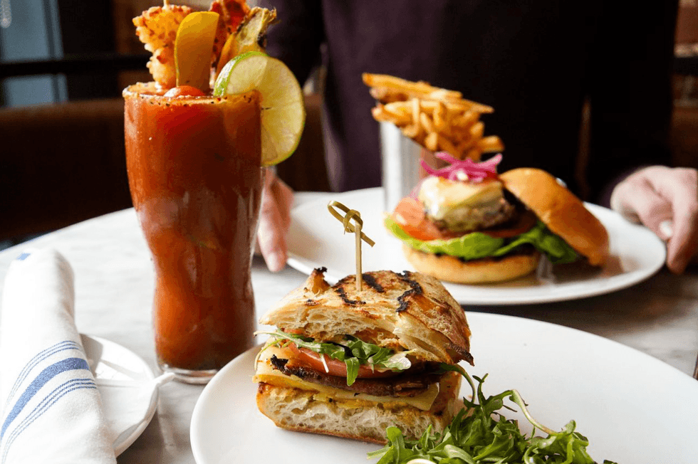 Sandwich, burger, and Bloody Mary on a table with fries in background.