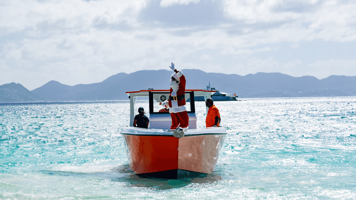 Santa waving on a red boat in tropical waters with mountains in the background.