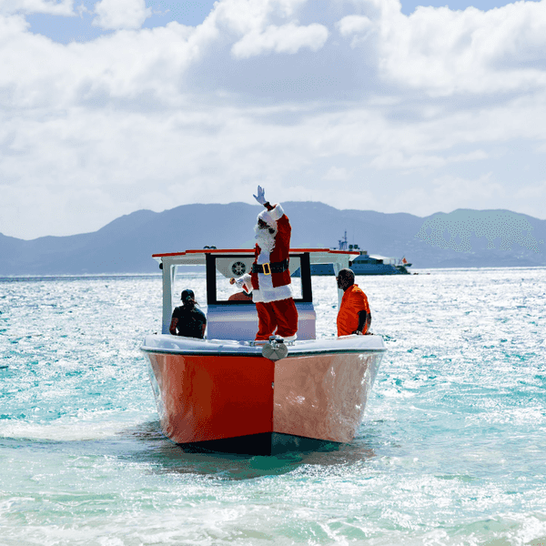 Santa waving on a red boat in tropical waters with mountains in the background.