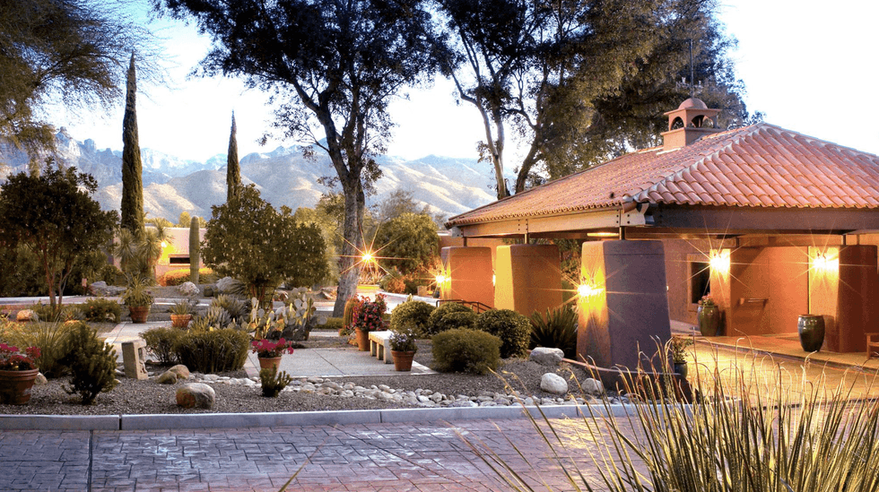 Scenic courtyard with trees, plants, a tiled-roof building, and mountains in the background.