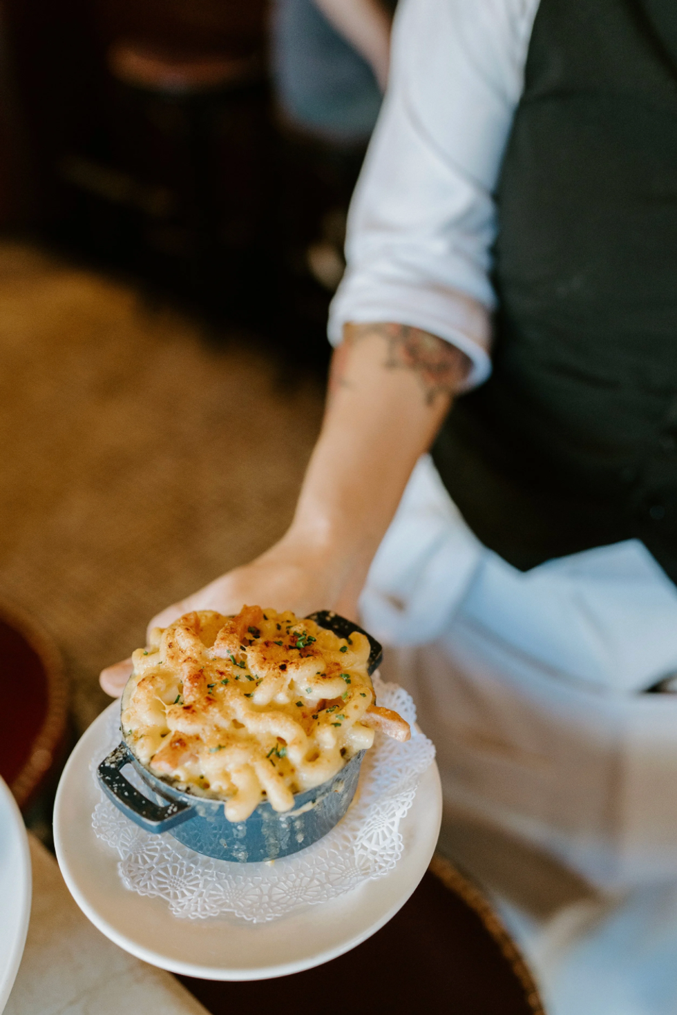Server presenting creamy mac and cheese in a small skillet on a doily-lined plate.
