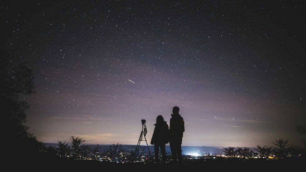 Silhouettes stargazing with a telescope under a clear, starry night sky.