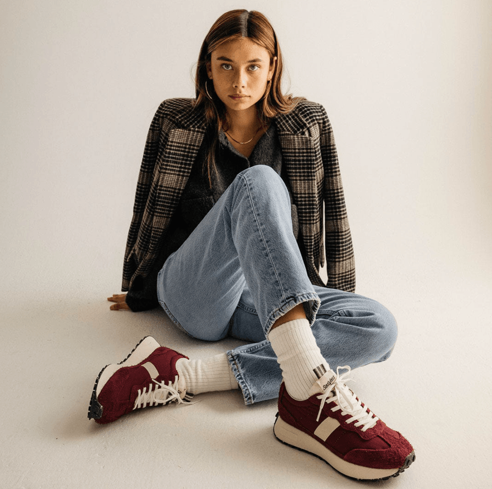 Sitting woman in plaid jacket, jeans, red sneakers, and white socks against a plain background.