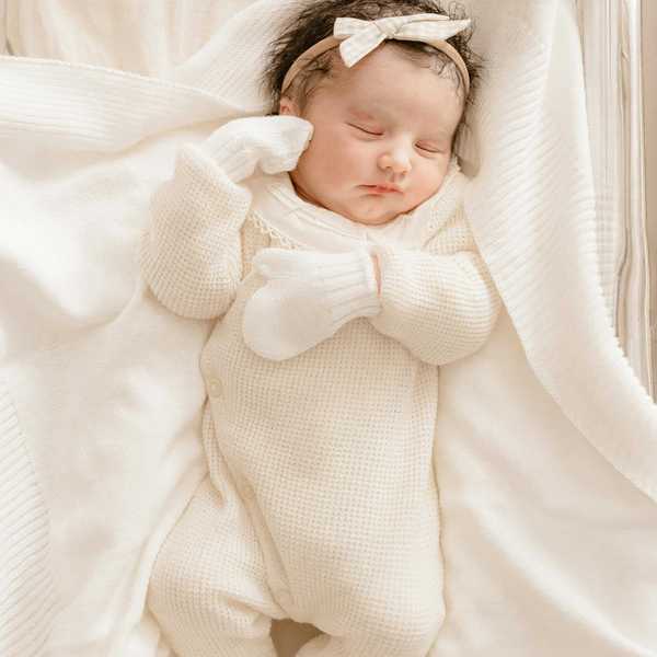 Sleeping baby in cream outfit with a bow headband, lying on a soft, white blanket.
