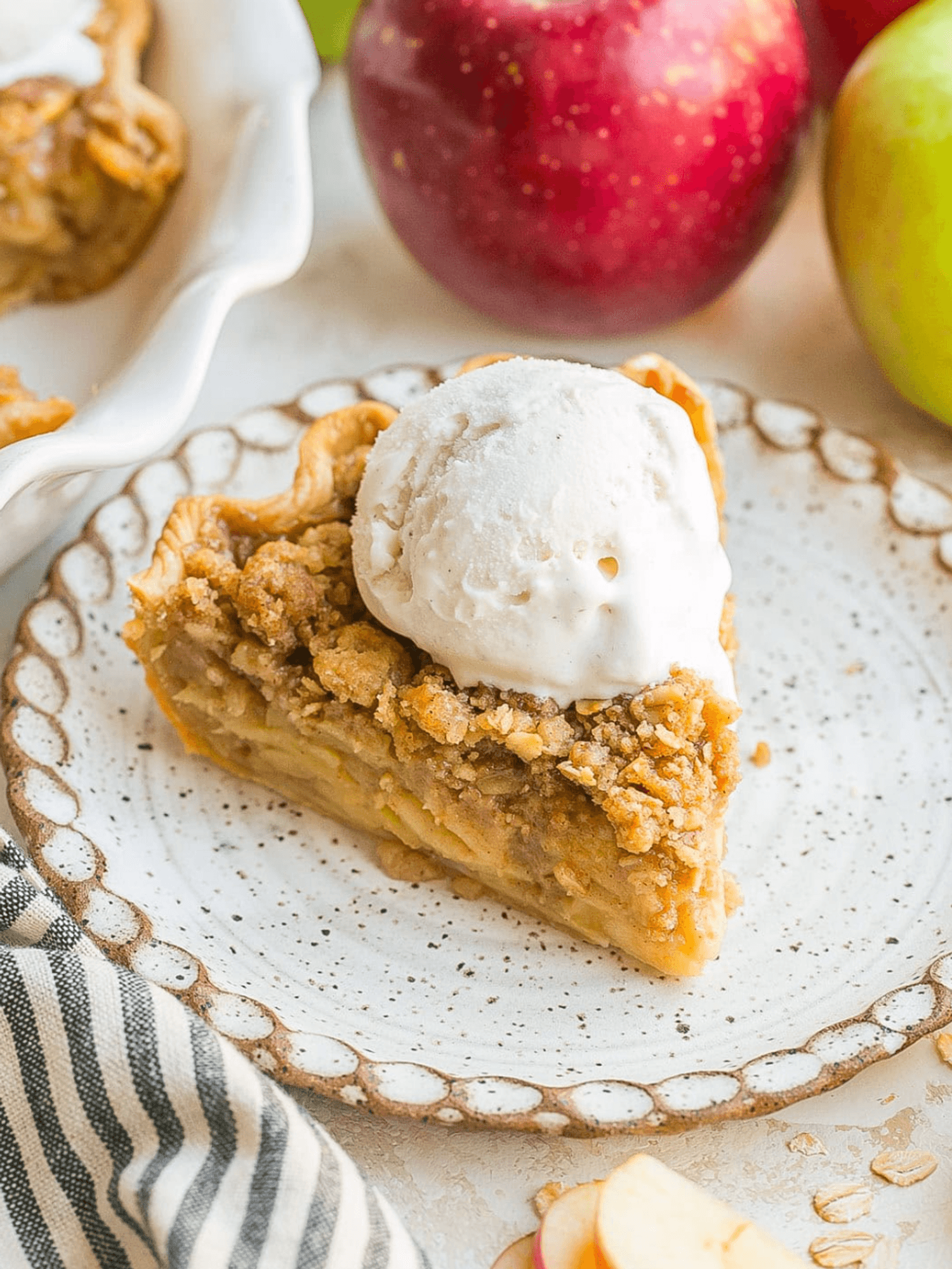 Slice of apple pie with ice cream on a plate, apples in the background.