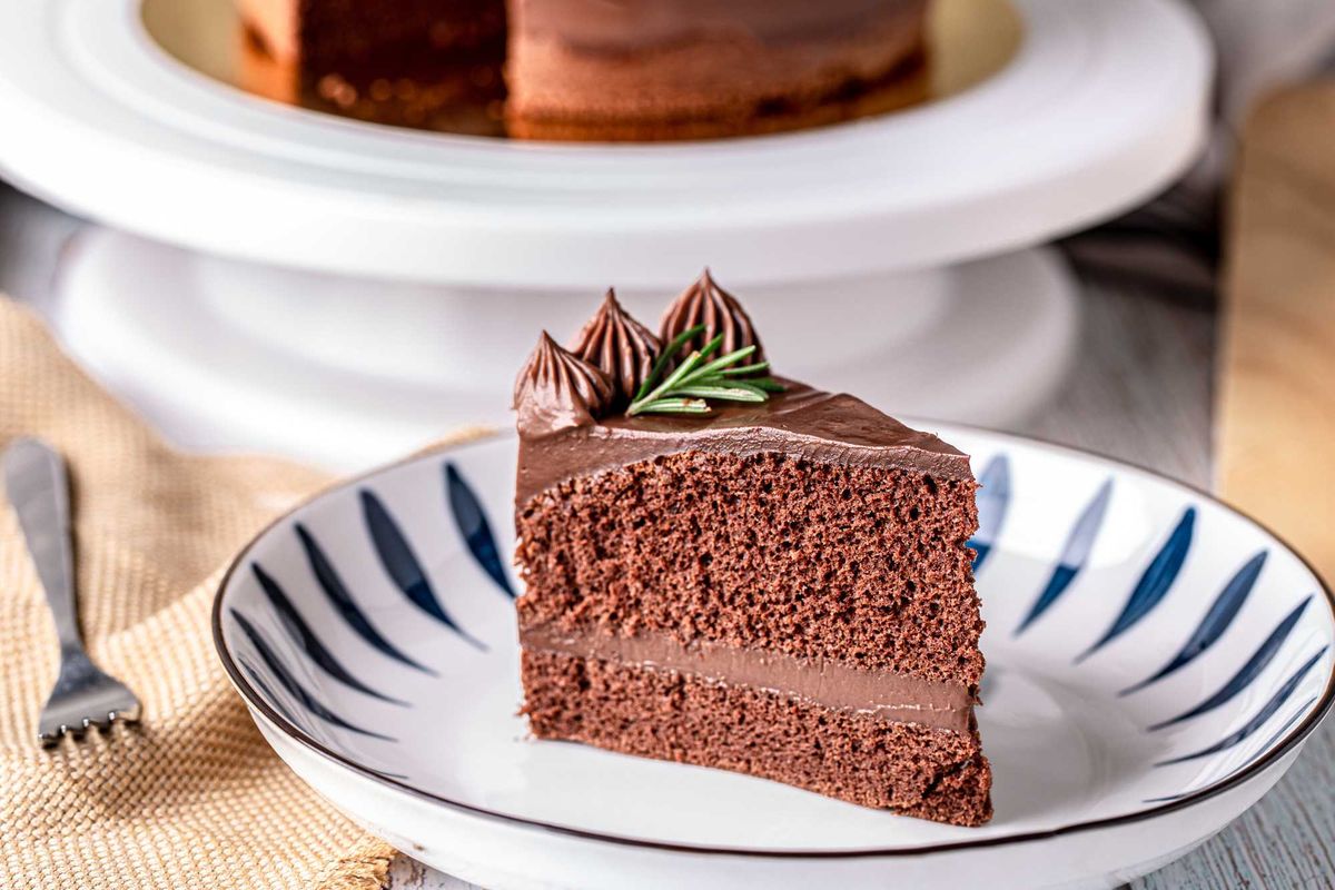 Slice of chocolate cake with frosting and rosemary garnish on a patterned plate.