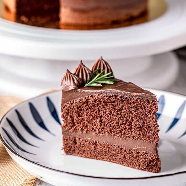 Slice of chocolate cake with frosting and rosemary garnish on a patterned plate.