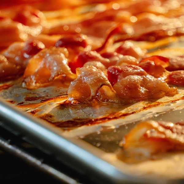 Slices of bacon sizzling and browning on a baking sheet in the oven.