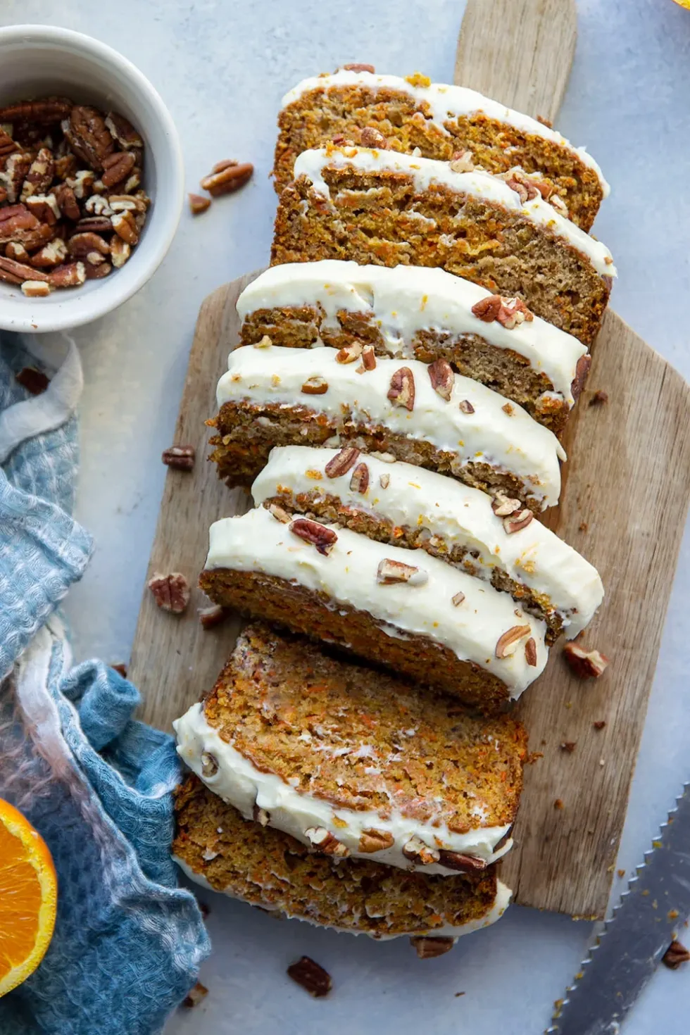 Slices of carrot cake banana bread, with white orange cream cheese frosting, rest on top of a wooden board with a bowl of pecans in the background.