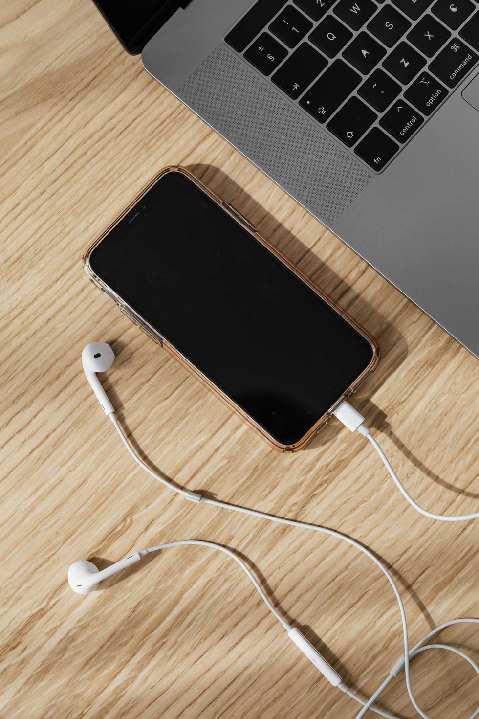 Smartphone with white earbuds connected, next to a laptop on a wooden surface.