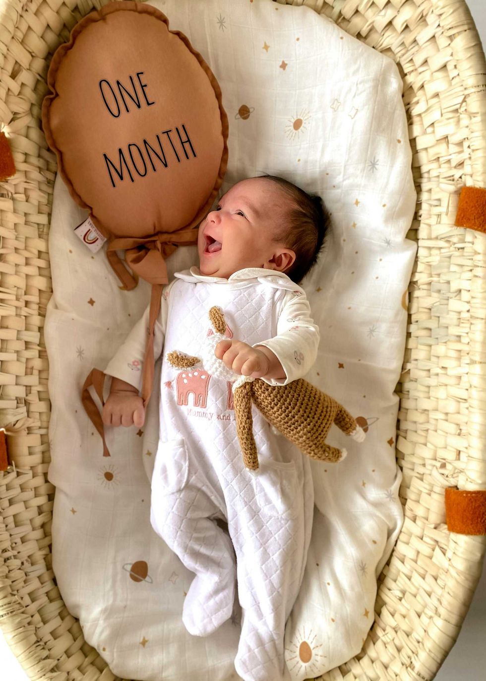 Smiling baby in a cradle with a "One Month" pillow and a knitted toy.