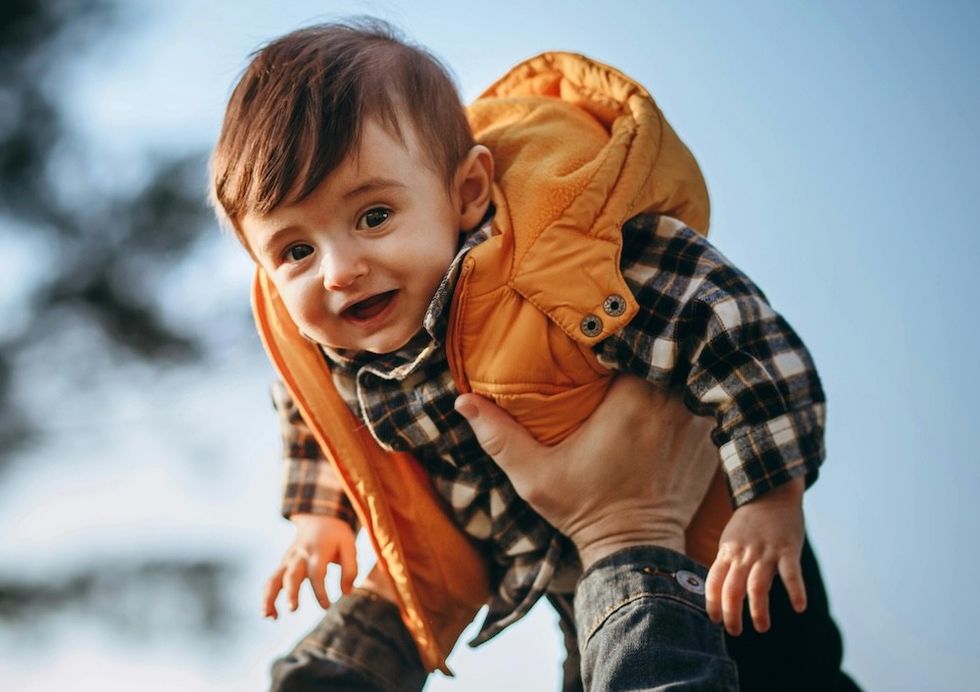Smiling baby in orange vest, lifted outdoors on a sunny day.