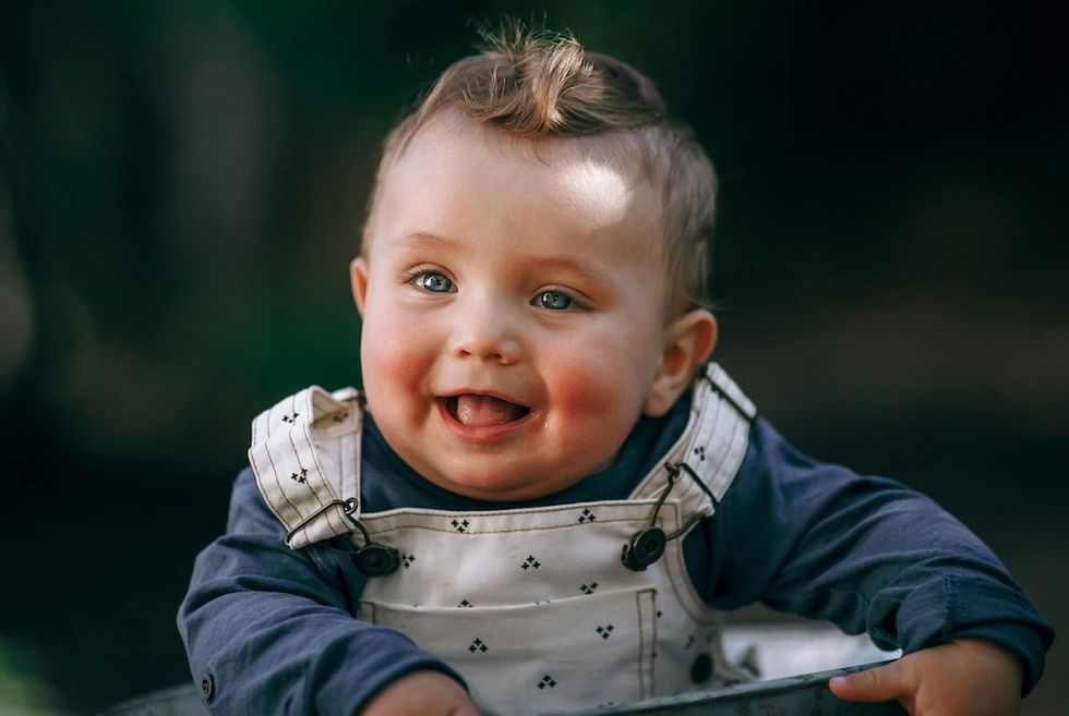Smiling baby in overalls with tiny patterns and a blue shirt, in soft natural light.