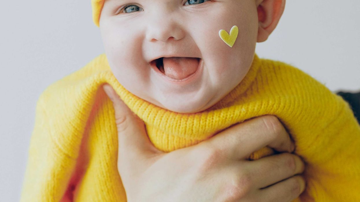 Smiling baby in yellow outfit and hat, held by an adult's hands, with heart sticker on cheek.