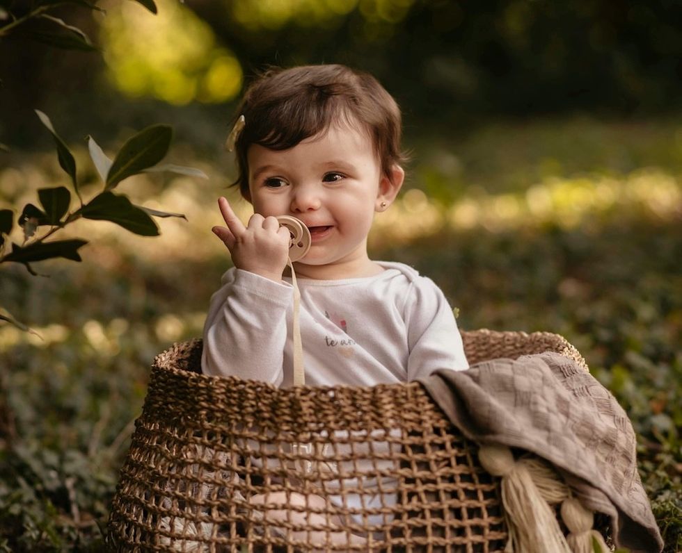 Smiling baby with pacifier sits in a wicker basket outdoors, surrounded by greenery.