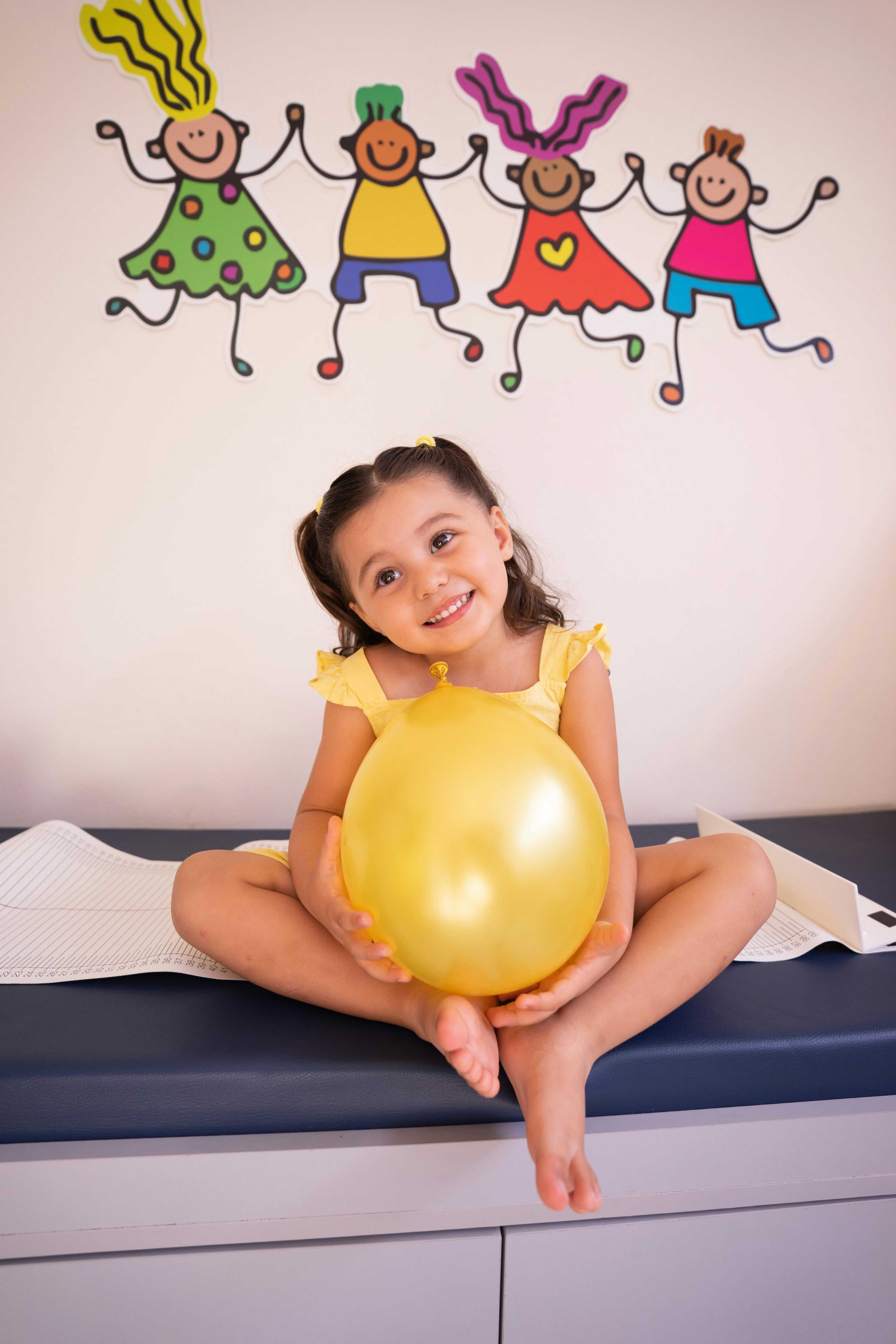 Smiling child in yellow holding balloon, sitting by colorful wall decor.