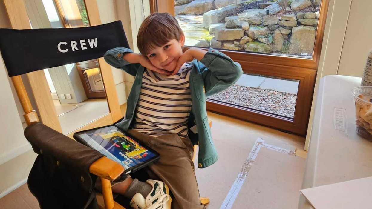 Smiling child on a director's chair with a tablet, indoors by a window.