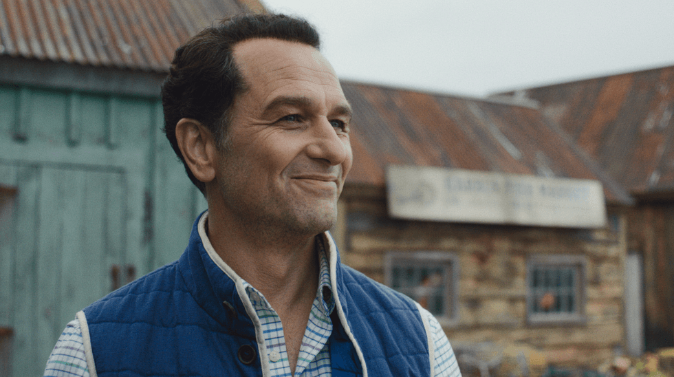 Smiling man in blue vest outside a rustic building.