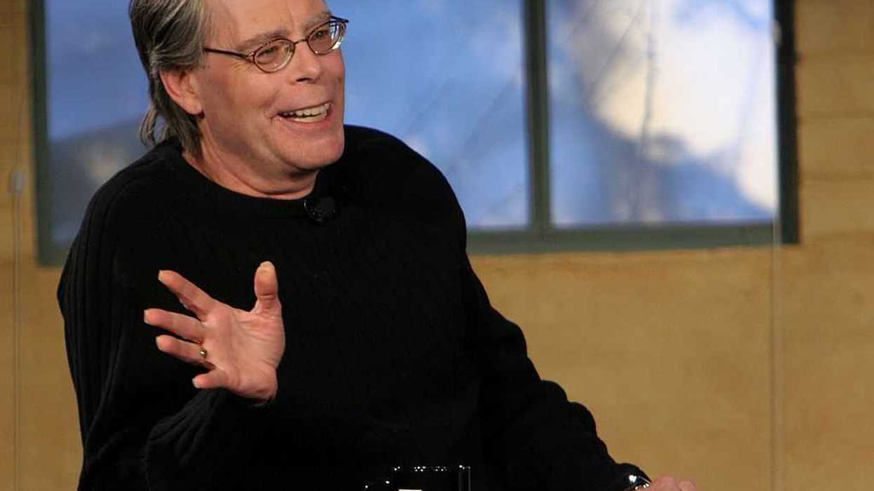 Smiling man in glasses gesturing, seated at a table with a black mug, in front of a window.