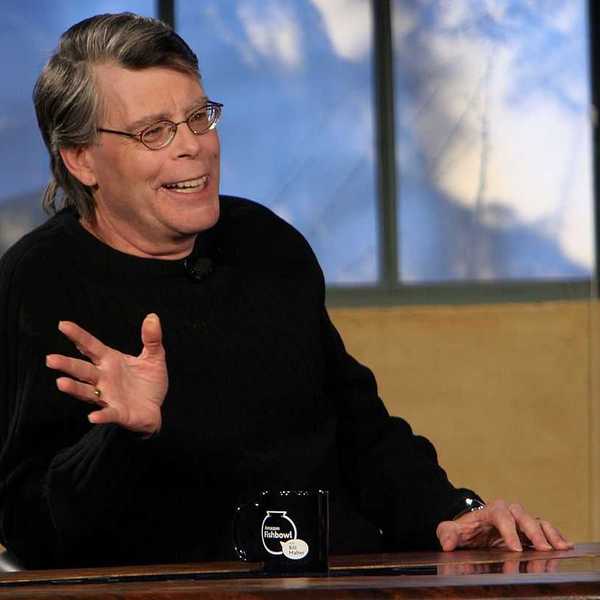 Smiling man in glasses gesturing, seated at a table with a black mug, in front of a window.