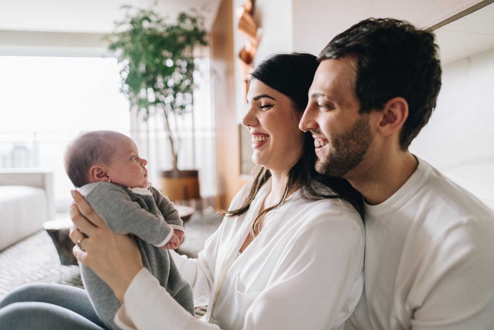 Smiling parents sitting together, holding a baby indoors.