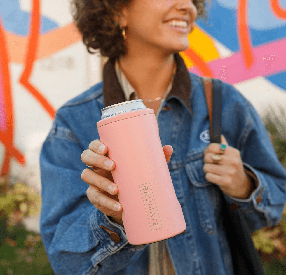 Smiling person holding a pink Br\u00fcMate can cooler against a colorful mural background.