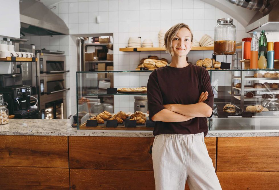 Smiling person in a bakery with arms crossed, surrounded by pastries and baked goods.