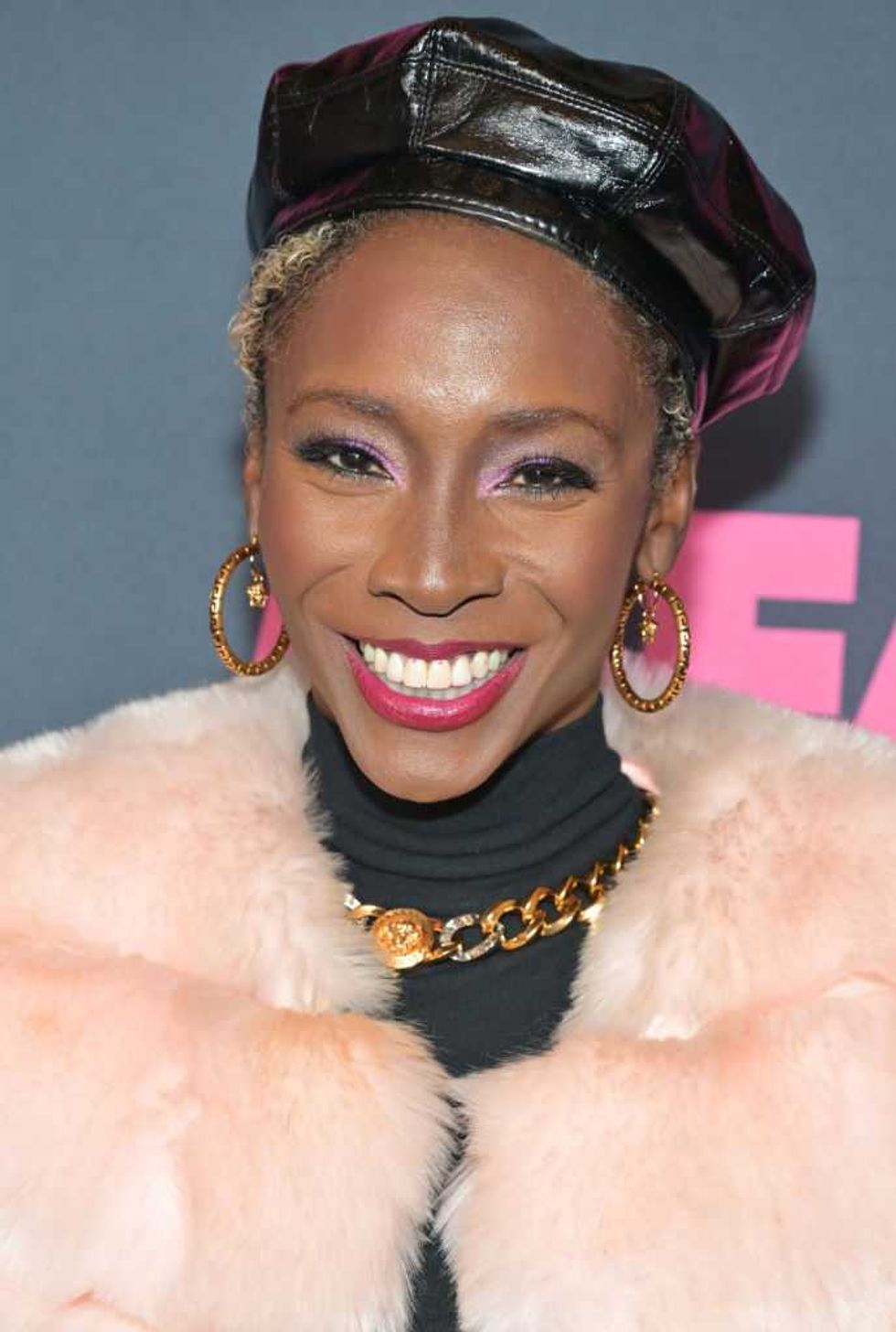 Smiling person in a leather beret, hoop earrings, and a fluffy pale pink coat.