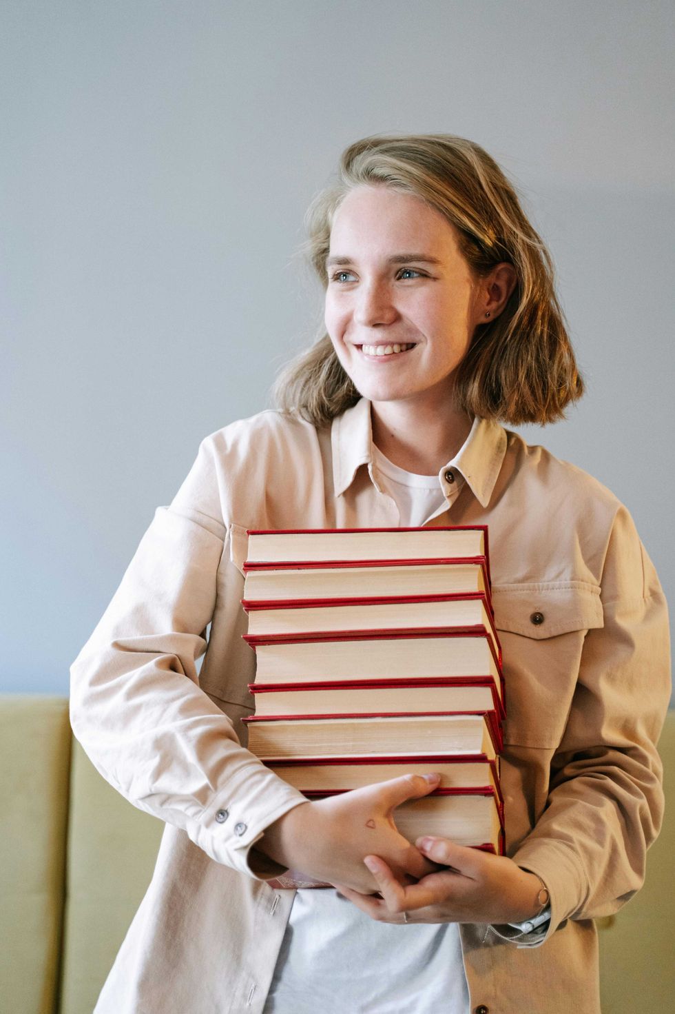 Smiling person in beige shirt holding a stack of books with red covers.