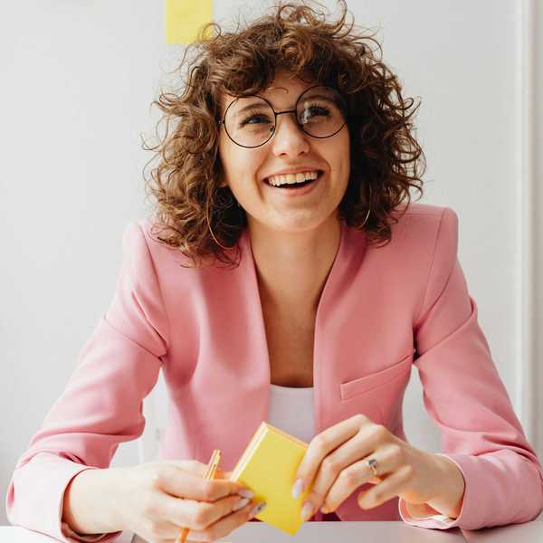Smiling person in glasses and a pink blazer sits at a desk with sticky notes.