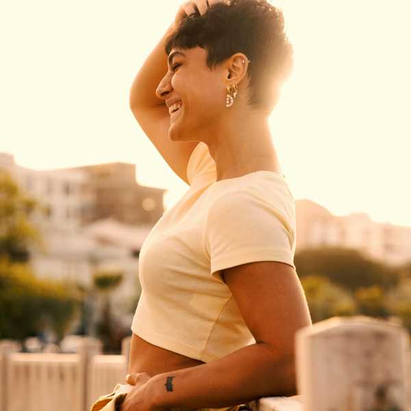 Smiling person in sunlight, wearing a yellow crop top, hand in hair, with cityscape background.