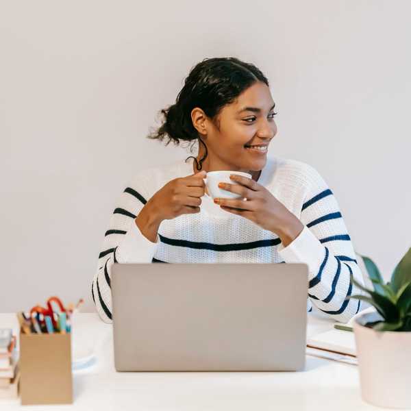 Smiling person with a coffee cup, sitting at a desk with a laptop and potted plant.