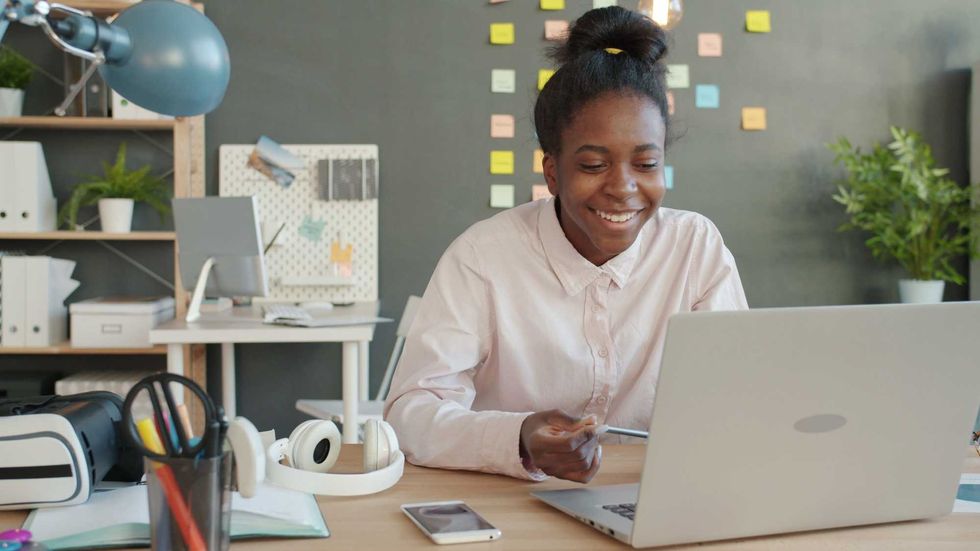 Smiling person works on a laptop at a desk with colorful sticky notes on the wall.