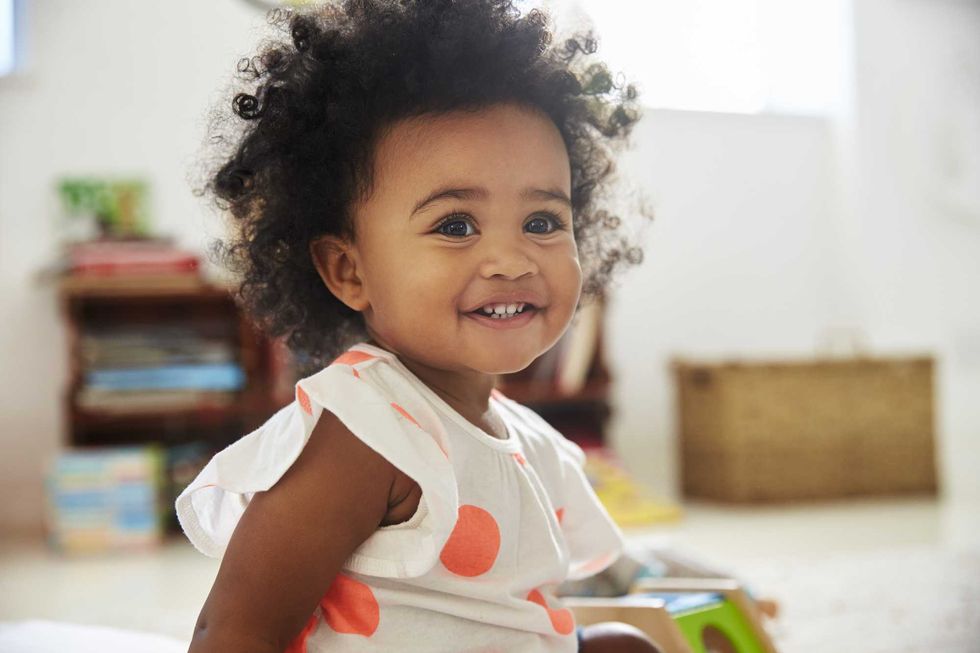 Smiling toddler with curly hair in a polka dot dress, sitting indoors.