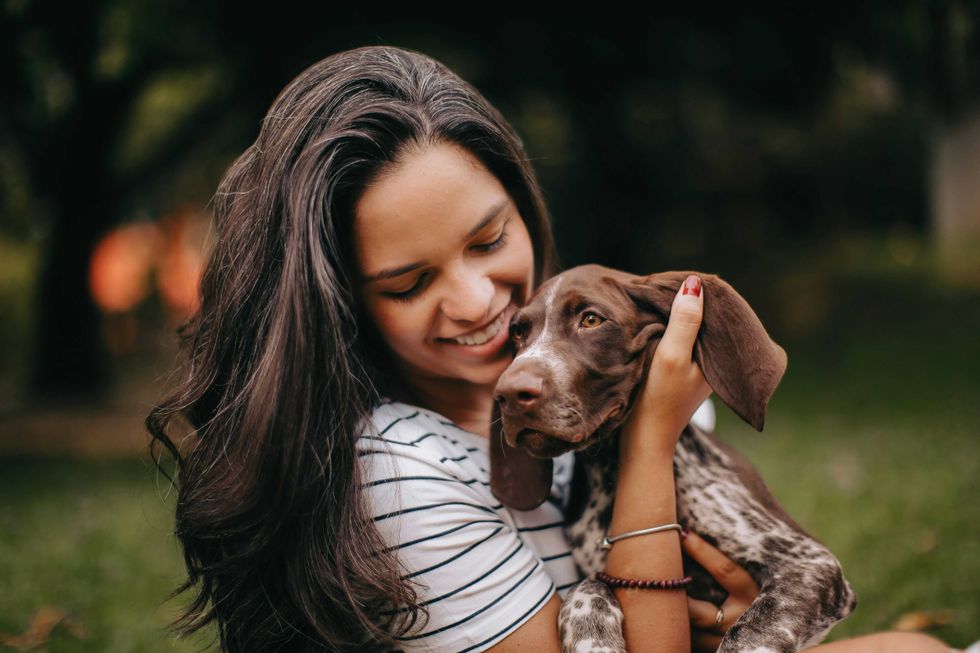 Smiling woman cuddles a brown and white puppy in a park setting.