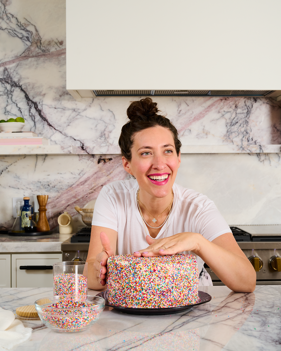 Smiling woman decorates a sprinkle-covered cake in a marble kitchen.