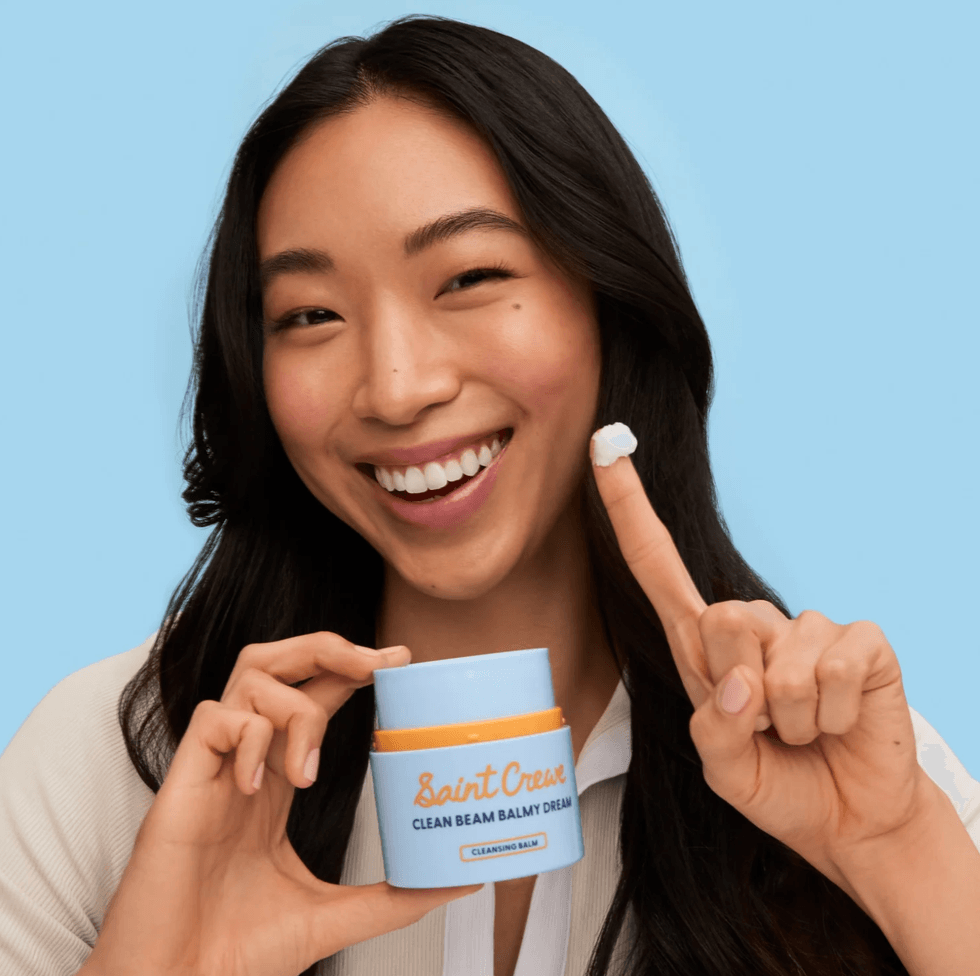 Smiling woman holding skincare balm, finger with product, blue background.