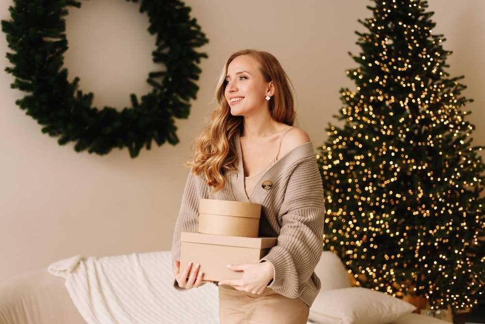 Smiling woman holds gift boxes near decorated Christmas tree and wreath.