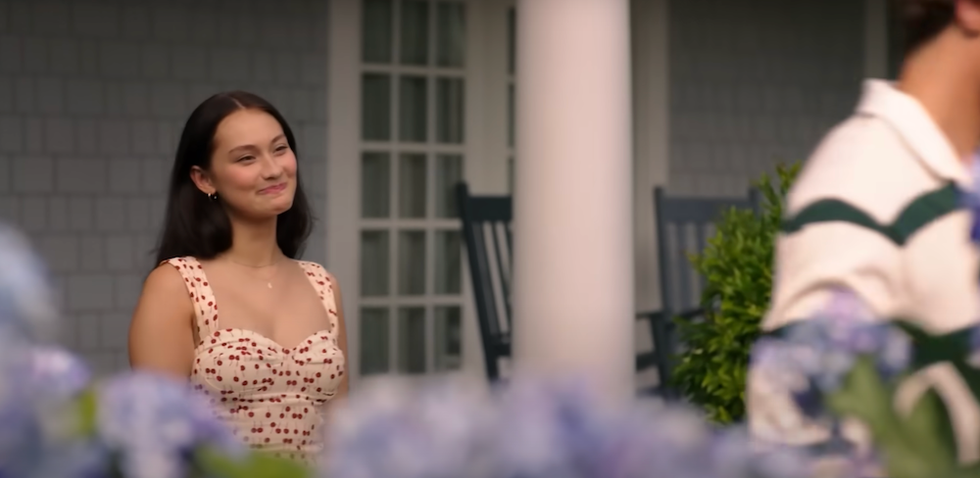 Smiling woman in a floral dress stands outside near hydrangeas and rocking chairs. lola tung as Belly conklin the summer i turned pretty season 3 episode 4