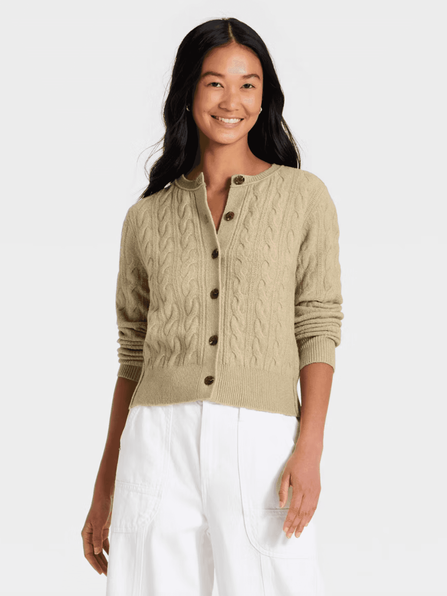Smiling woman in beige cable knit cardigan and white pants, standing against a plain background.