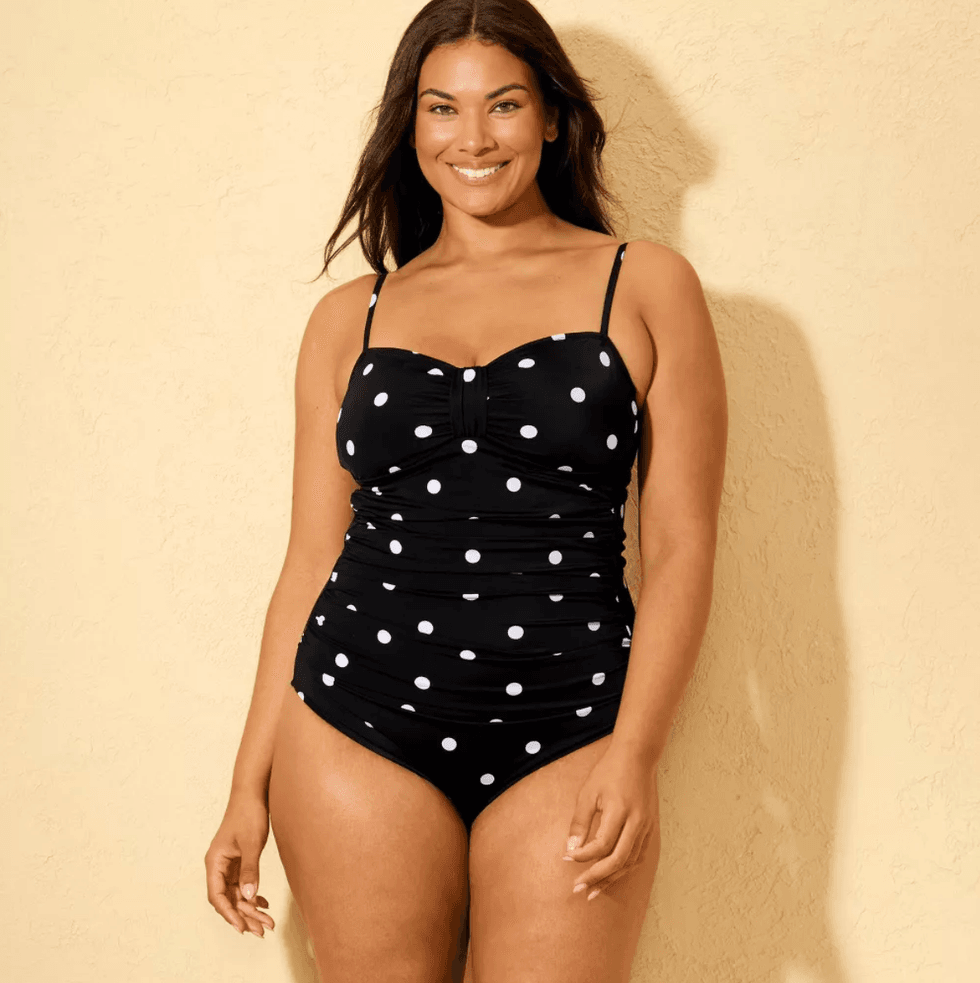 Smiling woman in black polka dot one-piece swimsuit against a beige background.