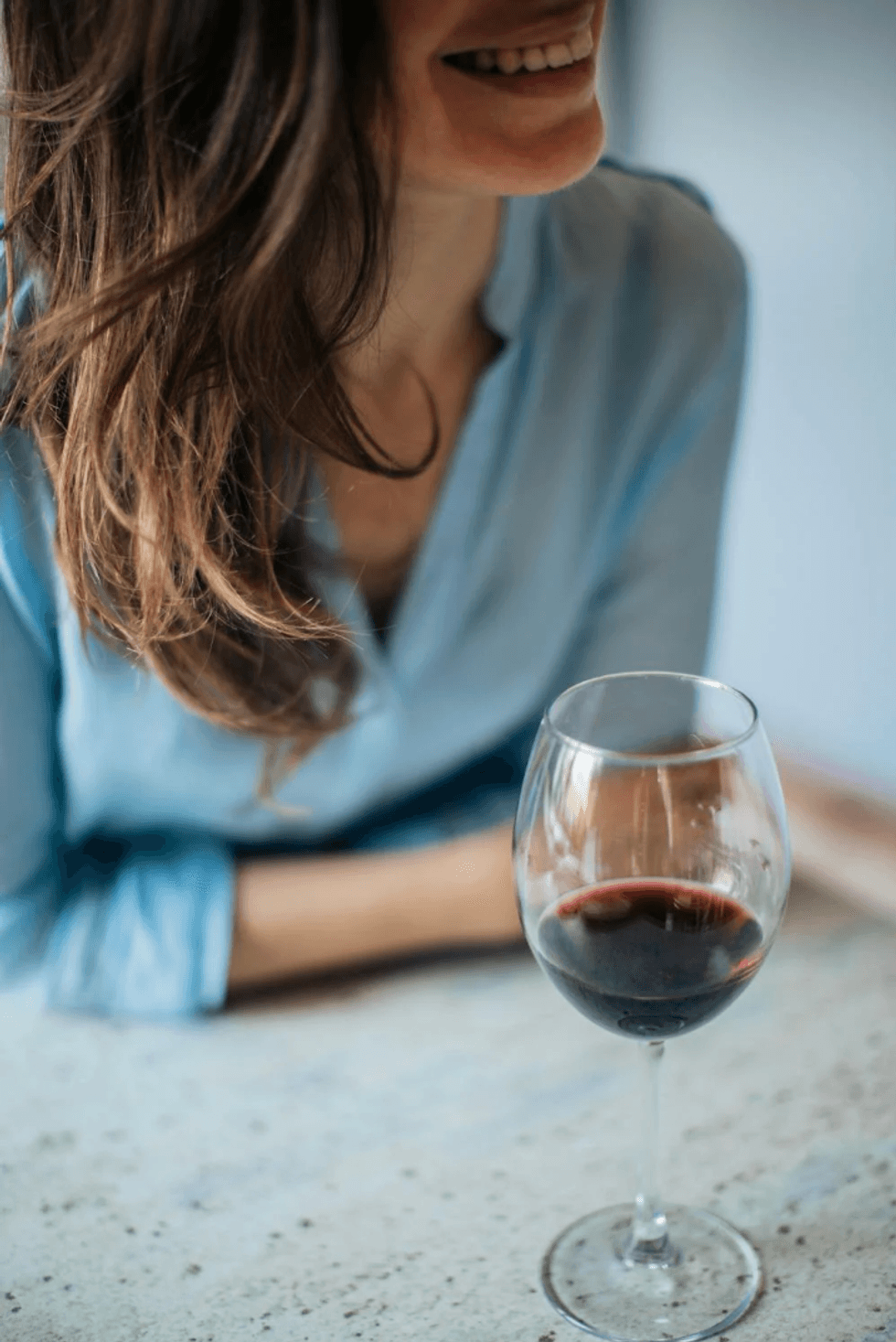 Smiling woman in blue shirt with a glass of red wine on a table.