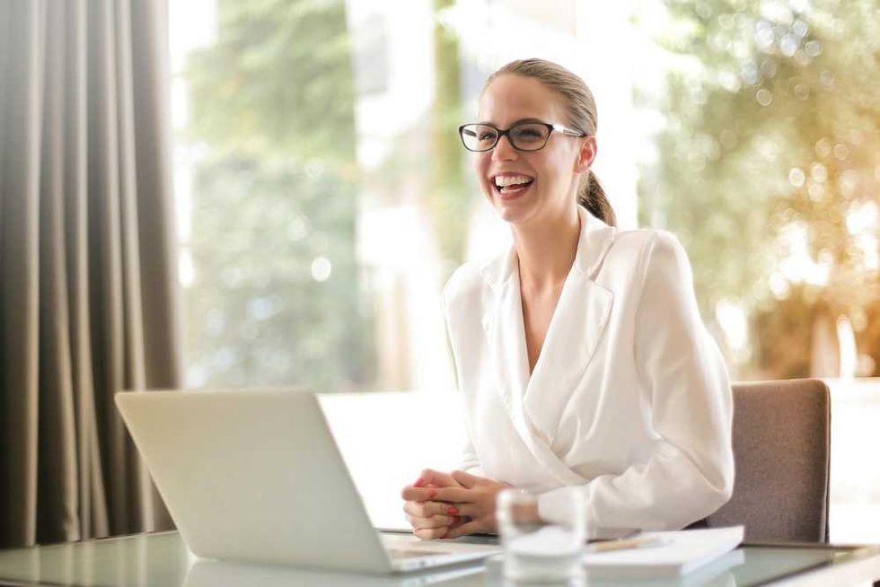 Smiling woman in glasses and white blazer working on a laptop at a desk.