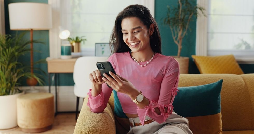 Smiling woman in pink shirt using smartphone on a cozy sofa.