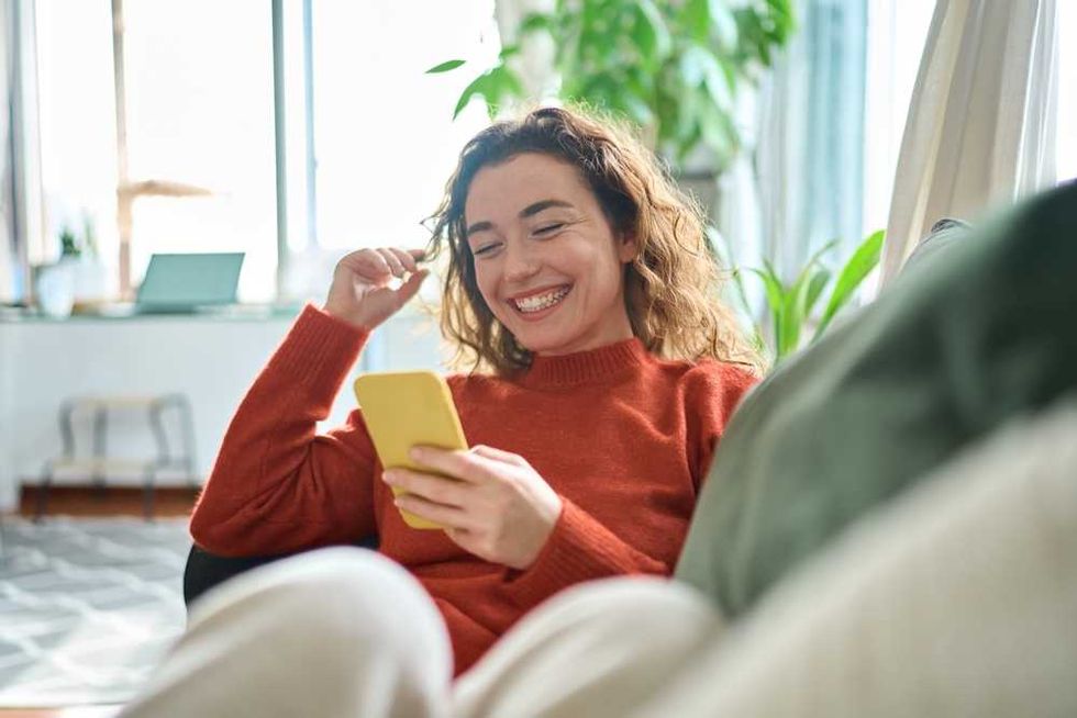 Smiling woman in red sweater using a yellow smartphone, sitting on a couch at home.