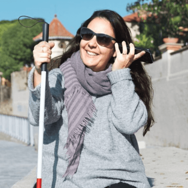 Smiling woman in sunglasses with cane, sitting outdoors, adjusts scarf.