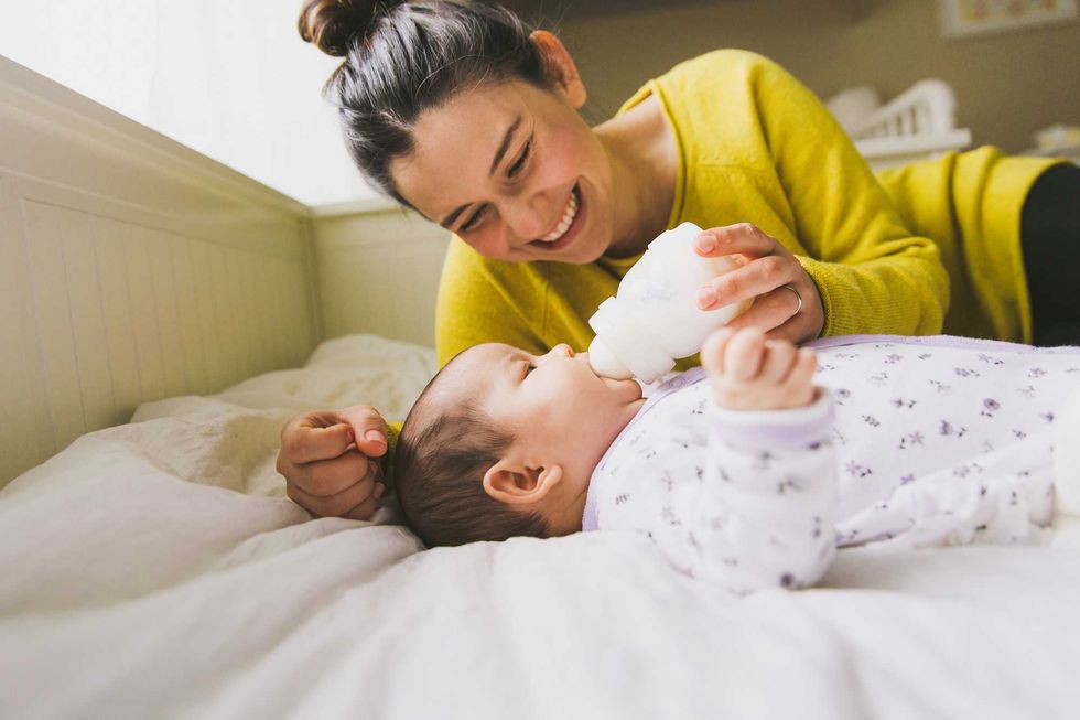 Smiling woman in yellow feeding a baby lying on a bed with a bottle.