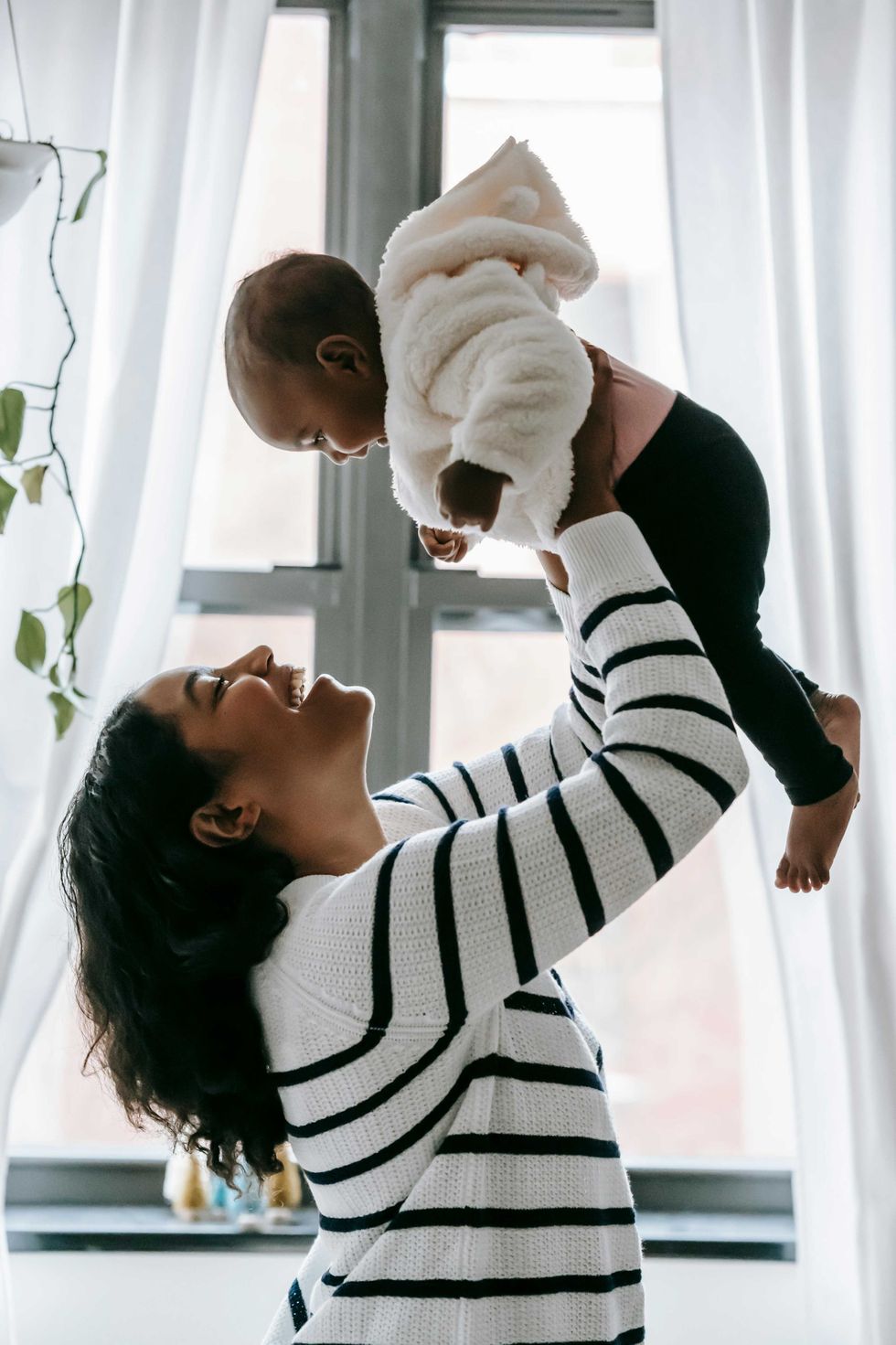 Smiling woman lifting baby in a cozy, light-filled room.